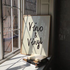 Puede incluir: Un cartel enmarcado con las palabras "Vino & Vinyl" en escritura negra. El cartel est&aacute; apoyado contra una ventana, con una pila de libros delante. El marco es de color madera clara.