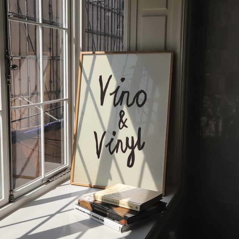 Puede incluir: Un cartel enmarcado con las palabras "Vino & Vinyl" en escritura negra. El cartel est&aacute; apoyado contra una ventana, con una pila de libros delante. El marco es de color madera clara.