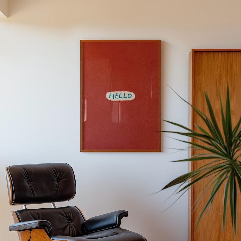 Puede incluir: Una obra de arte enmarcada con un fondo rojo y la palabra "HELLO" en un &oacute;valo blanco y azul. El marco es de color marr&oacute;n c&aacute;lido. Una silla de cuero negro y una planta est&aacute;n en primer plano.