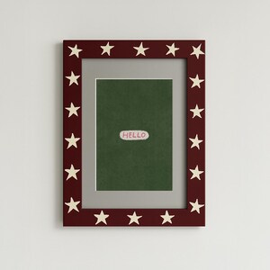 Puede incluir: Un marco de fotos rectangular con un borde rojo oscuro adornado con estrellas blancas. El marco contiene un fondo verde con una peque&ntilde;a pegatina ovalada blanca que dice "HELLO". El marco est&aacute; dise&ntilde;ado para mostrar fotos u obras de arte.