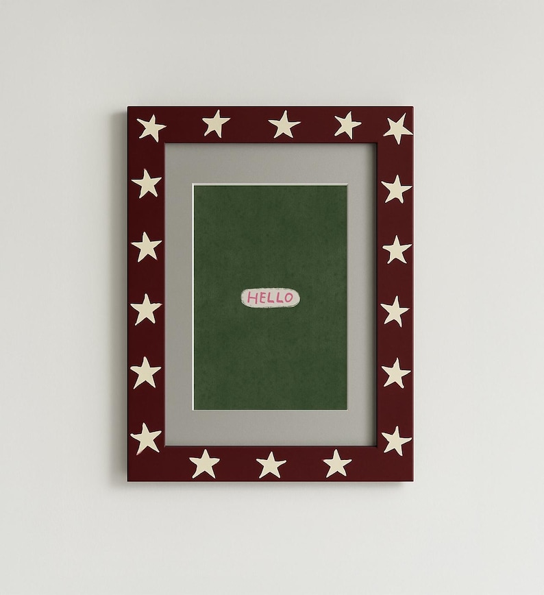 Puede incluir: Un marco de fotos rectangular con un borde rojo oscuro adornado con estrellas blancas. El marco contiene un fondo verde con una peque&ntilde;a pegatina ovalada blanca que dice "HELLO". El marco est&aacute; dise&ntilde;ado para mostrar fotos u obras de arte.