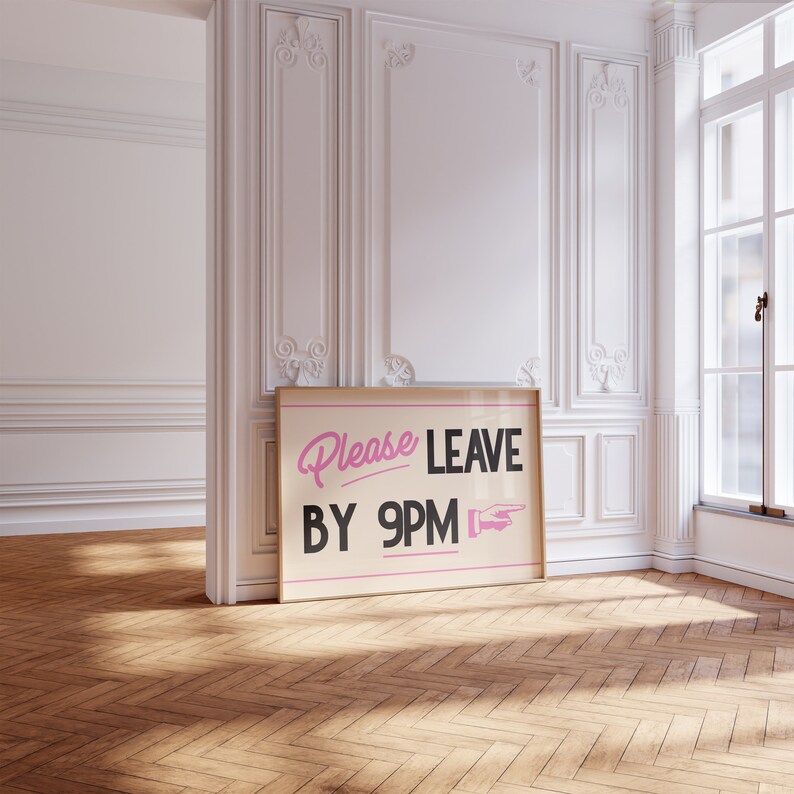 May include: A framed poster with the text "Please LEAVE BY 9PM" in pink and black. The poster is leaning against a white wall in a room with a wooden floor. The room has a window and decorative molding.