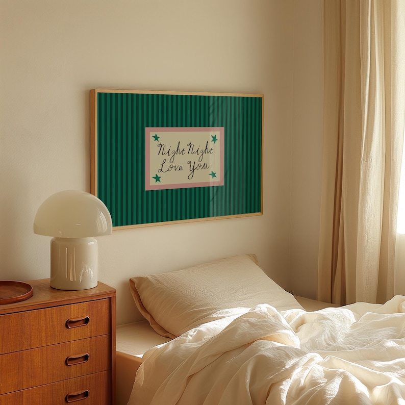 K&ouml;nnte beinhalten: Ein gerahmtes Wandbild mit gr&uuml;n gestreiftem Hintergrund und einem rosa Rechteck in der Mitte. Der Text "Night Night Love You" steht in Schreibschrift innerhalb des rosa Rechtecks, mit gr&uuml;nen Sternen. Eine wei&szlig;e Pilzlampe steht auf einem Nachttisch aus Holz.