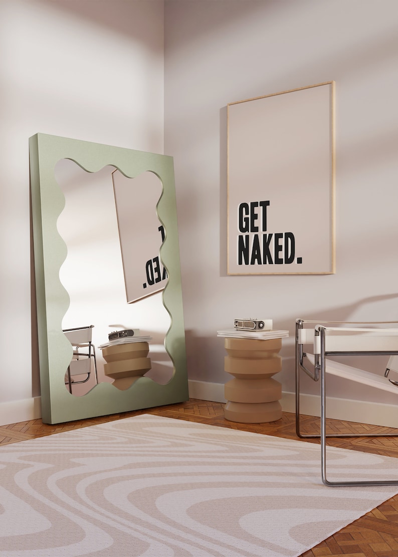May include: A large floor mirror with a sage green wavy frame, a beige cylindrical side table, and a rectangular wall art piece featuring the black text GET NAKED. The mirror reflects the room, including the side table and the wall art.