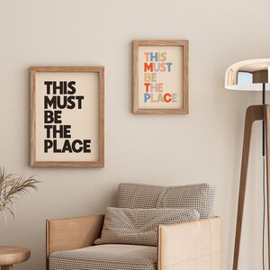Puede incluir: Dos impresiones enmarcadas con el texto "This must be the place" en blanco y negro y letras de colores, colgadas en una pared beige sobre un sill&oacute;n marr&oacute;n con estampado a cuadros y una l&aacute;mpara de pie de madera con pantalla plateada.