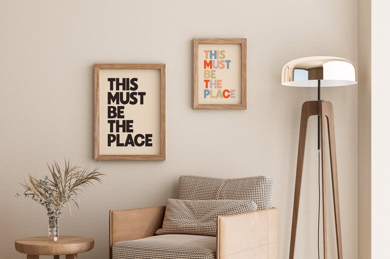 Puede incluir: Dos impresiones enmarcadas con el texto "This must be the place" en blanco y negro y letras de colores, colgadas en una pared beige sobre un sill&oacute;n marr&oacute;n con estampado a cuadros y una l&aacute;mpara de pie de madera con pantalla plateada.