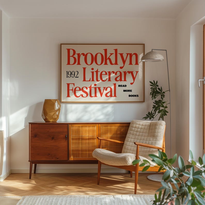 May include: A room with a mid-century modern aesthetic. A wooden cabinet with a geometric vase sits below a framed poster that reads "Brooklyn Literary Festival 1992". A beige armchair and a floor lamp complete the scene.