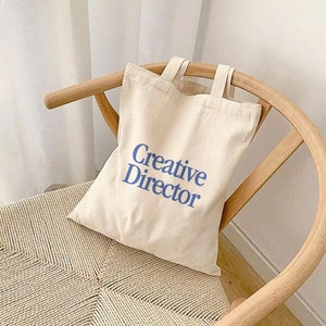 May include: A white canvas tote bag with blue text that reads "Creative Director". The bag is sitting on a light brown wicker chair.