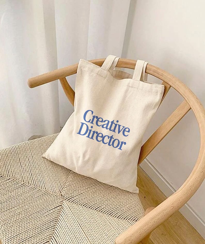 May include: A white canvas tote bag with blue text that reads "Creative Director". The bag is sitting on a light brown wicker chair.