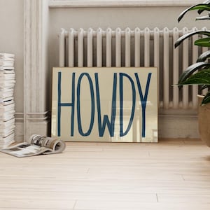 Puede incluir: Una impresi&oacute;n enmarcada con la palabra "HOWDY" escrita en una fuente grande, negrita y azul sobre un fondo beige.