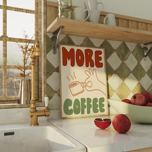 May include: A retro-style poster with the text "MORE COFFEE" in colorful, stylized lettering. A cartoon hand holds a steaming cup of coffee. The poster has a cream background with green and orange accents.