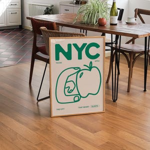 May include: A framed print with a green and white design. The print features the word "NYC" in large green letters, with the words "The Big Apple" and "The City That Never Sleeps" in smaller green letters below. The print also includes a green outline of a hand holding an apple.