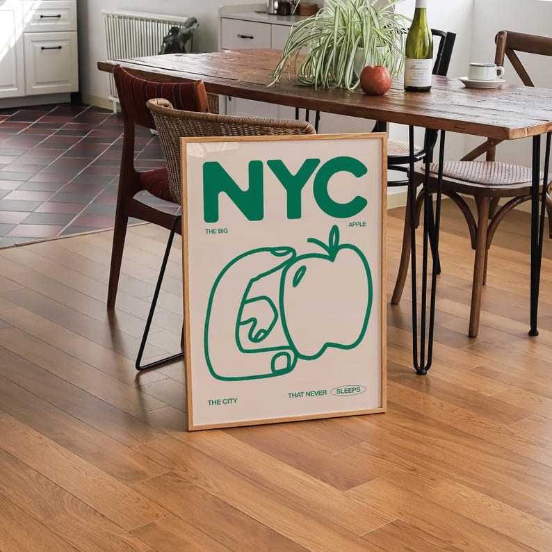 May include: A framed print with a green and white design. The print features the word "NYC" in large green letters, with the words "The Big Apple" and "The City That Never Sleeps" in smaller green letters below. The print also includes a green outline of a hand holding an apple.