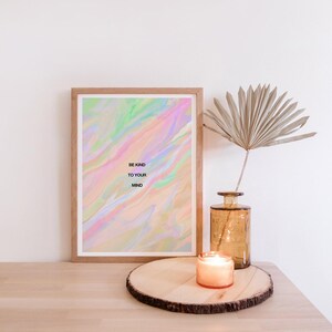 May include: A framed print with the text "BE KIND TO YOUR MIND" against a pastel, swirled background. A lit candle sits on a wooden slab, next to a vase with a dried palm leaf. The scene is set on a light-colored wooden surface.