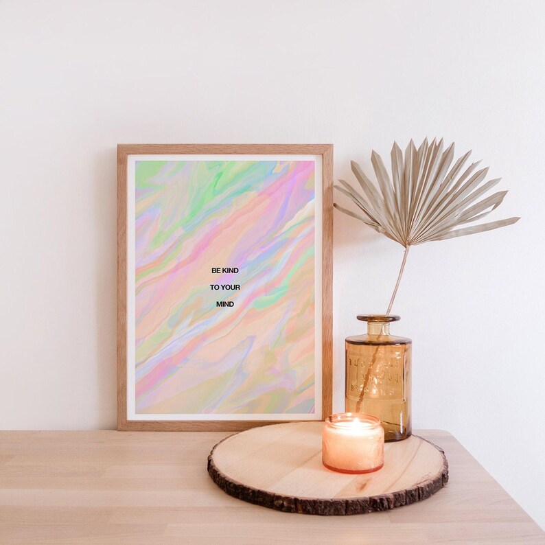 May include: A framed print with the text "BE KIND TO YOUR MIND" against a pastel, swirled background. A lit candle sits on a wooden slab, next to a vase with a dried palm leaf. The scene is set on a light-colored wooden surface.