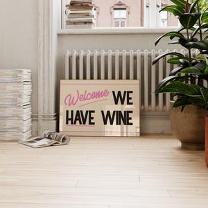 May include: A beige sign with the text "Welcome We Have Wine" in black lettering. The word "Welcome" is written in pink cursive.