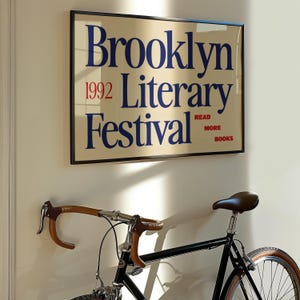 May include: A framed poster for the Brooklyn Literary Festival 1992, with blue text on a cream background, hangs above a black bicycle. The bicycle has brown handlebars and a brown leather seat.