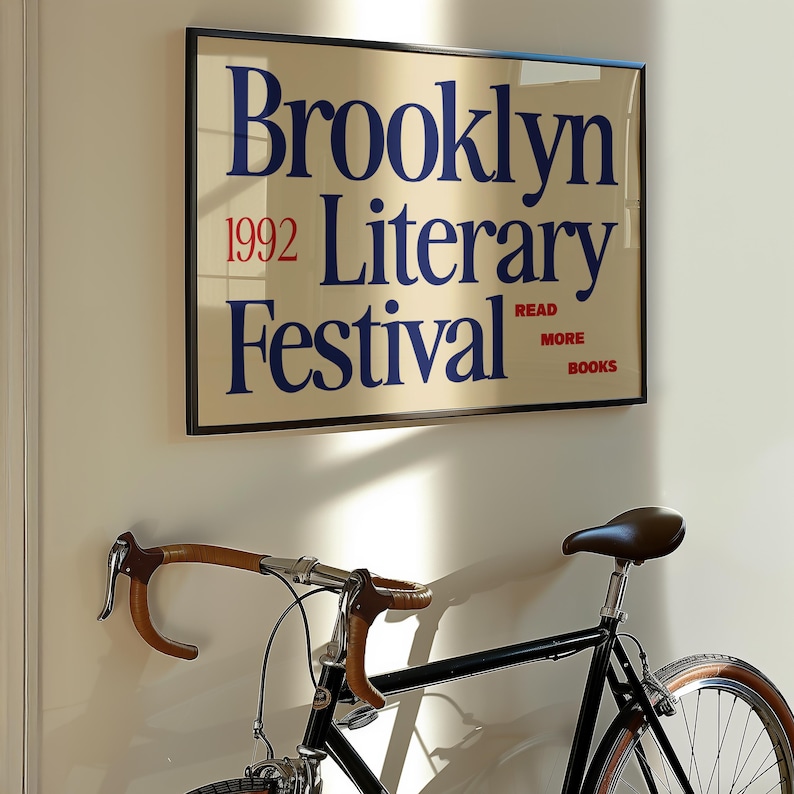 May include: A framed poster for the Brooklyn Literary Festival 1992, with blue text on a cream background, hangs above a black bicycle. The bicycle has brown handlebars and a brown leather seat.
