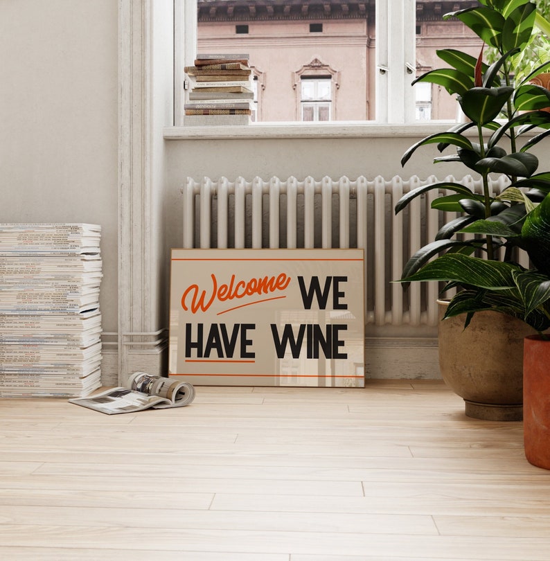 May include: A beige sign with black text that reads "Welcome We Have Wine" in a retro font. The text "Welcome" is in orange. The sign is hanging on a white wall in a room with a window and a radiator.