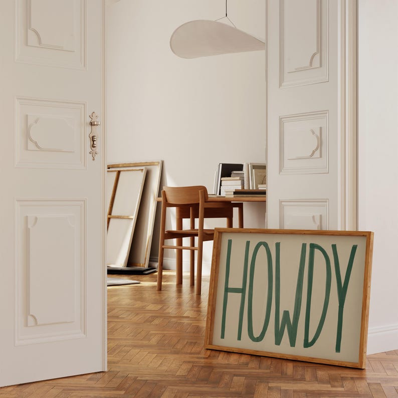 Puede incluir: Una impresi&oacute;n sobre lienzo enmarcada con la palabra "HOWDY" escrita en verde sobre un fondo beige. El marco es de madera natural y tiene un aspecto r&uacute;stico. La impresi&oacute;n se apoya contra una pared blanca en una habitaci&oacute;n con suelo de madera.