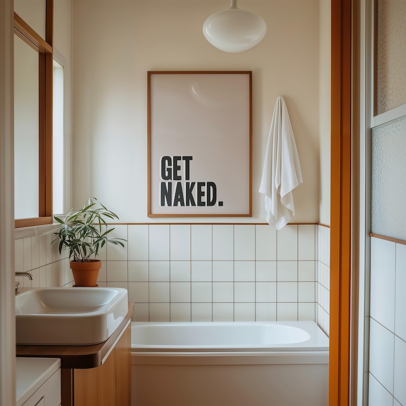 May include: A rectangular wall art print in a thin wooden frame featuring the black text GET NAKED. centered on a light beige background. The print is displayed on a wall above a white bathtub with white square tiles and a white towel hanging nearby.