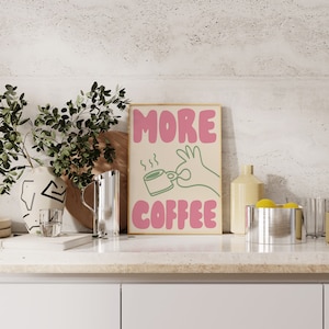May include: A framed print with the text "MORE COFFEE" in pink letters. A green hand is holding a steaming cup of coffee.