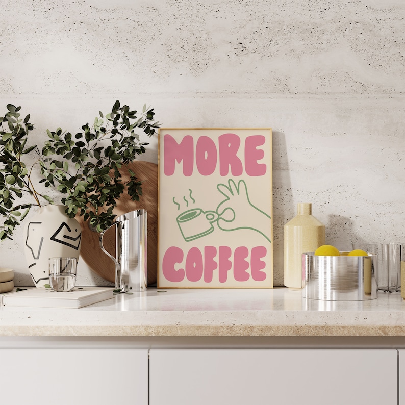 May include: A framed print with the text "MORE COFFEE" in pink letters. A green hand is holding a steaming cup of coffee.