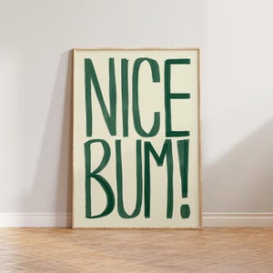 Può includere: Una stampa artistica rettangolare da parete con sfondo color crema chiaro e il testo NICE BUM! scritto a mano in verde scuro con uno stile a pennellate audaci. L'opera è racchiusa in una semplice cornice in legno di colore chiaro.