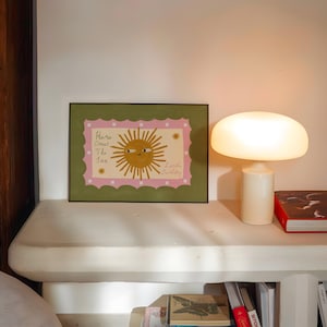 Puede incluir: Una impresi&oacute;n art&iacute;stica enmarcada con una ilustraci&oacute;n de un sol sonriente y el texto "Here Comes The Sun" y "Little Darling". La obra de arte tiene un borde rosa y verde. Una l&aacute;mpara con forma de hongo est&aacute; en un estante blanco.
