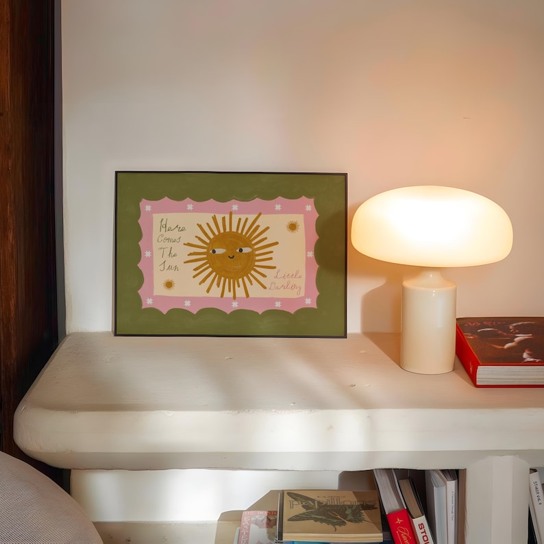Puede incluir: Una impresi&oacute;n art&iacute;stica enmarcada con una ilustraci&oacute;n de un sol sonriente y el texto "Here Comes The Sun" y "Little Darling". La obra de arte tiene un borde rosa y verde. Una l&aacute;mpara con forma de hongo est&aacute; en un estante blanco.
