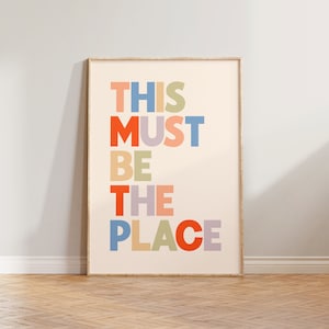 Puede incluir: Una impresi&oacute;n enmarcada con el texto "THIS MUST BE THE PLACE" en letras de colores sobre un fondo crema.
