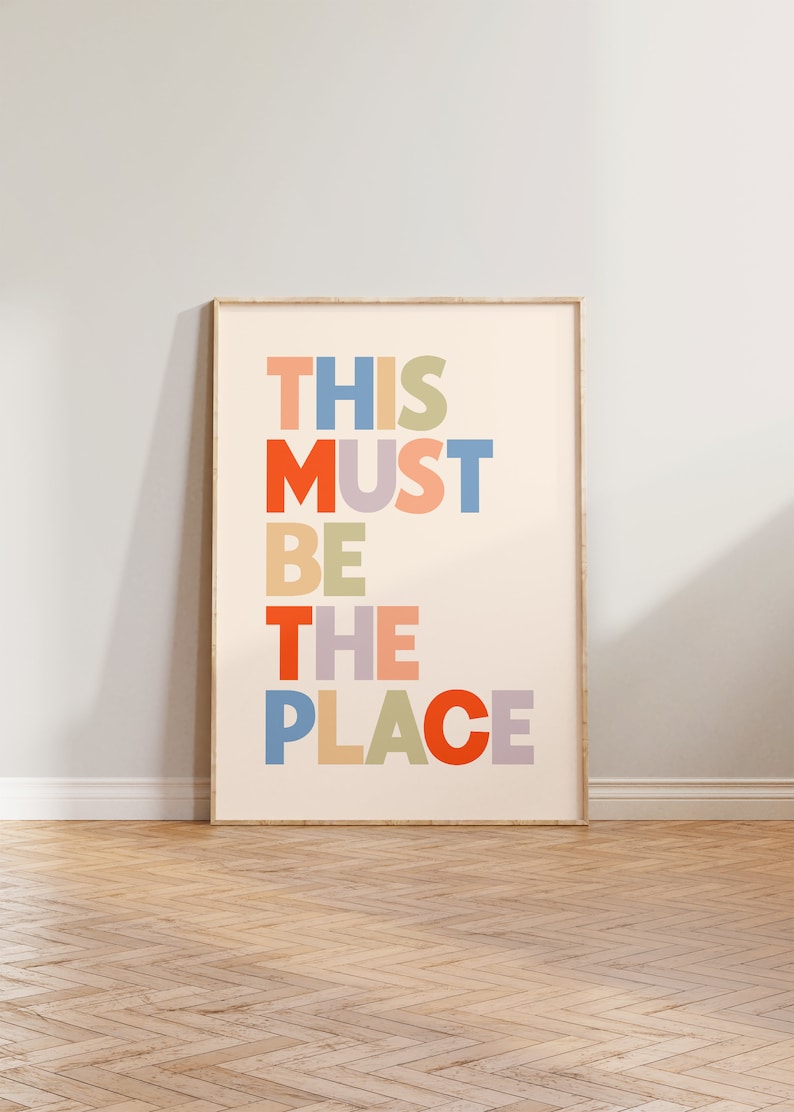 Puede incluir: Una impresi&oacute;n enmarcada con el texto "THIS MUST BE THE PLACE" en letras de colores sobre un fondo crema.