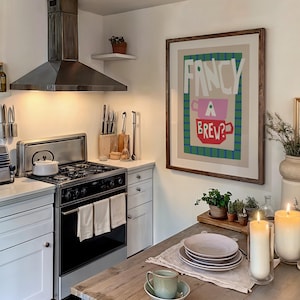 Op de afbeelding: Een keuken met witte kasten, een fornuis van roestvrij staal en een ingelijst afdruk op de muur met de tekst "Fancy a Brew?" De afdruk toont een roze en rode theekop met groene en blauwe strepen op de achtergrond.
