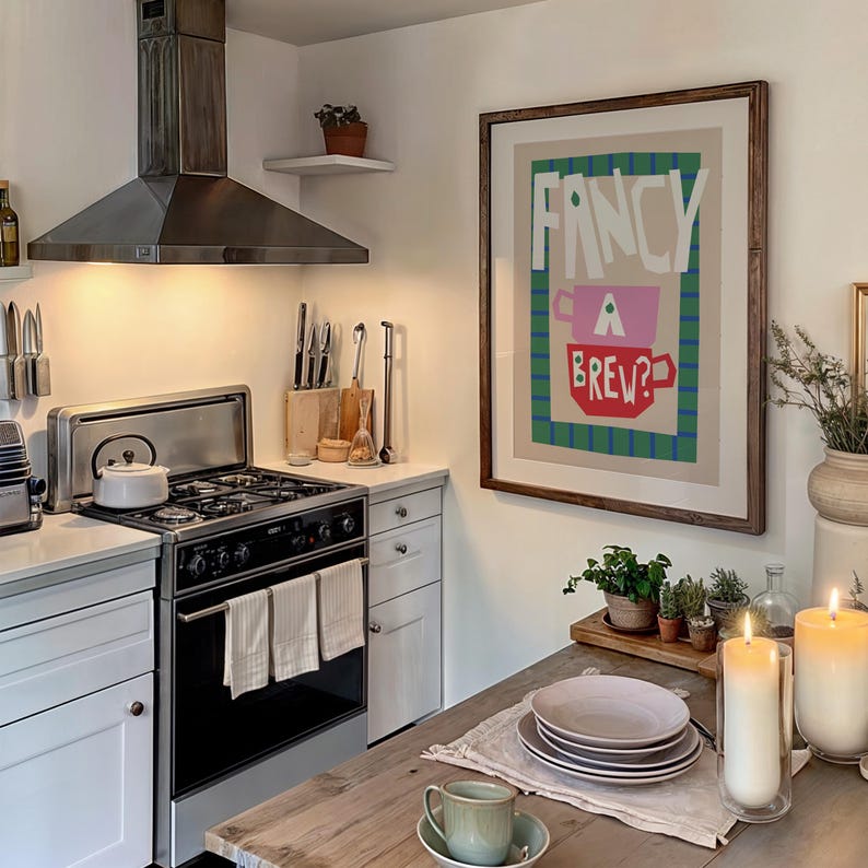 Op de afbeelding: Een keuken met witte kasten, een fornuis van roestvrij staal en een ingelijst afdruk op de muur met de tekst "Fancy a Brew?" De afdruk toont een roze en rode theekop met groene en blauwe strepen op de achtergrond.