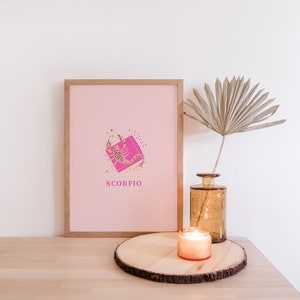 May include: A framed print featuring a light pink background with a central graphic of a scorpion over a bright pink square, surrounded by small gold stars and dots, with the word SCORPIO printed in pink below the illustration.