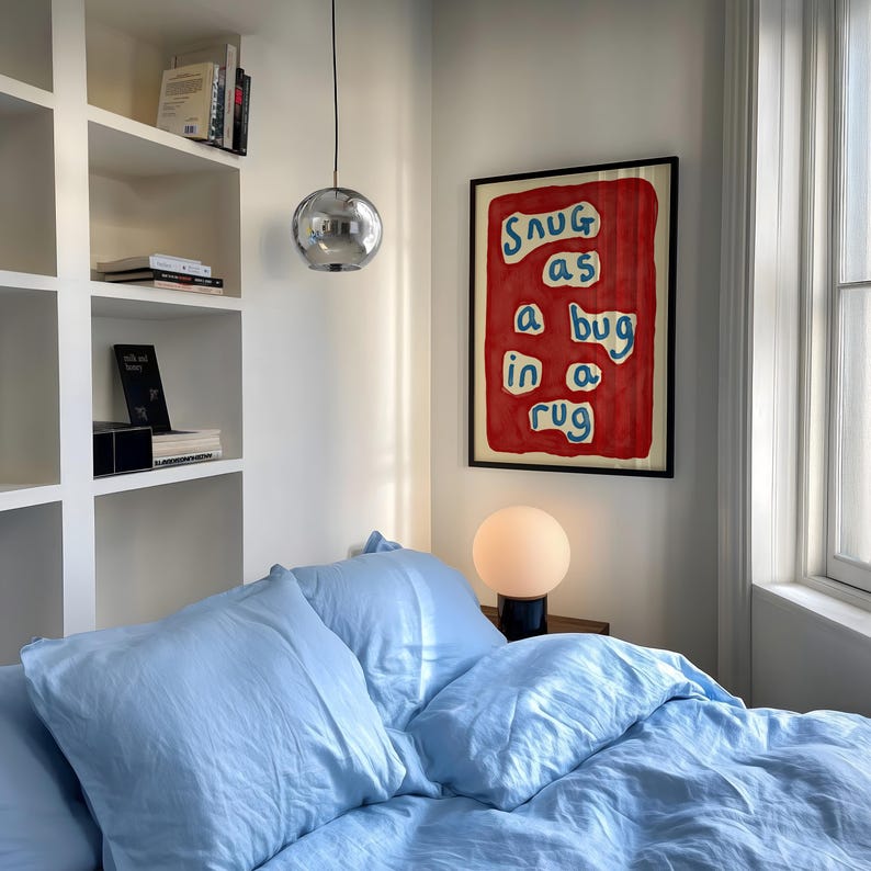 May include: A bedroom scene featuring a bed with light blue bedding and pillows. A framed artwork with the text "Snug as a bug in a rug" hangs on the wall. A spherical lamp and a chrome pendant light are also visible.