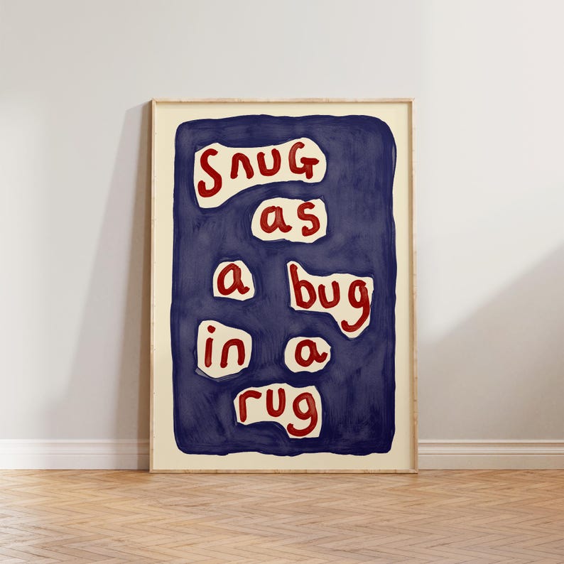 May include: A framed art print with the phrase "Snug as a bug in a rug" in red letters on a dark blue background. The artwork has a cream-colored border and is displayed on a light-colored wall.