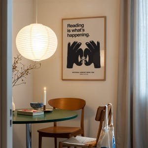 May include: A round, white paper lantern hangs above a small, green table. A framed poster with the text "Reading is what's happening" and a graphic of hands is on the wall. A lit candle and a wooden chair are also visible.