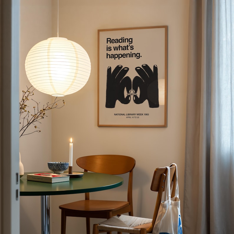May include: A round, white paper lantern hangs above a small, green table. A framed poster with the text "Reading is what's happening" and a graphic of hands is on the wall. A lit candle and a wooden chair are also visible.