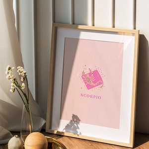 May include: A framed wall art print featuring a light pink background with a central illustration of a scorpion in a darker pink square. The word SCORPIO is printed in pink capital letters below the graphic, accented by small gold star icons.