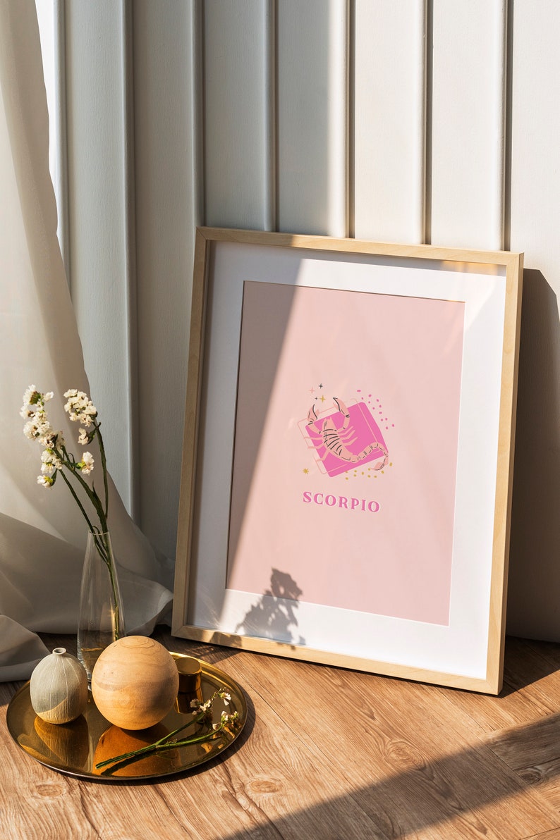 May include: A framed wall art print featuring a light pink background with a central illustration of a scorpion in a darker pink square. The word SCORPIO is printed in pink capital letters below the graphic, accented by small gold star icons.