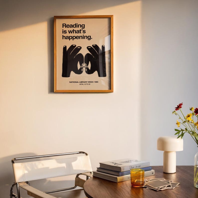 May include: A framed poster with the text "Reading is what's happening" and a graphic of two hands forming a book. The poster is in a wooden frame and is hanging on a white wall. A chair, books, and a lamp are in the foreground.