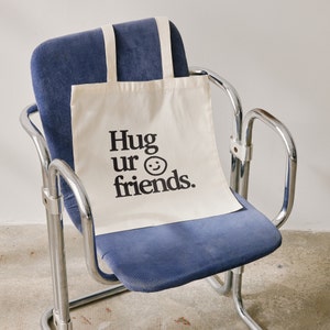 May include: A white canvas tote bag with black text that reads "Hug ur friends." and a black smiley face. The bag is sitting on a blue chair.