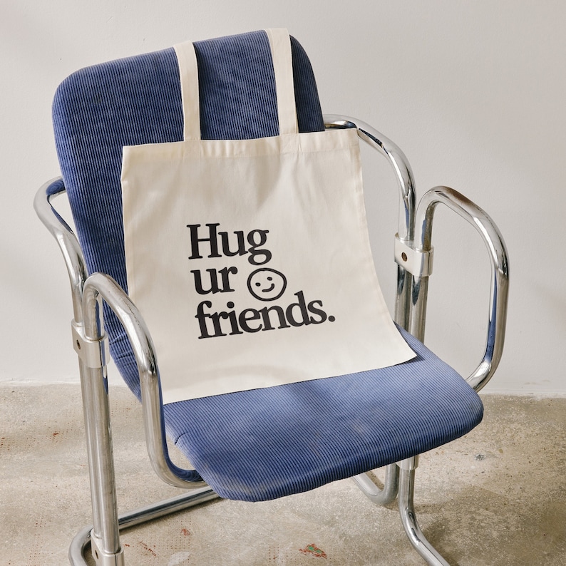 May include: A white canvas tote bag with black text that reads "Hug ur friends." and a black smiley face. The bag is sitting on a blue chair.