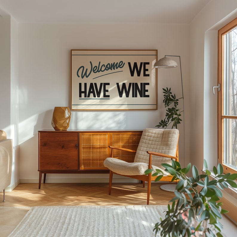 May include: A living room interior with a mid-century modern aesthetic. A wooden cabinet with a woven rattan door sits beneath a framed print that reads "Welcome We Have Wine". A beige armchair sits in front of the cabinet, and a potted plant is in the foreground.