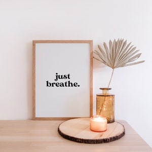 May include: A framed print with the text "just breathe." on a white background. The print is sitting on a wooden table next to a vase with a dried palm leaf and a lit candle.