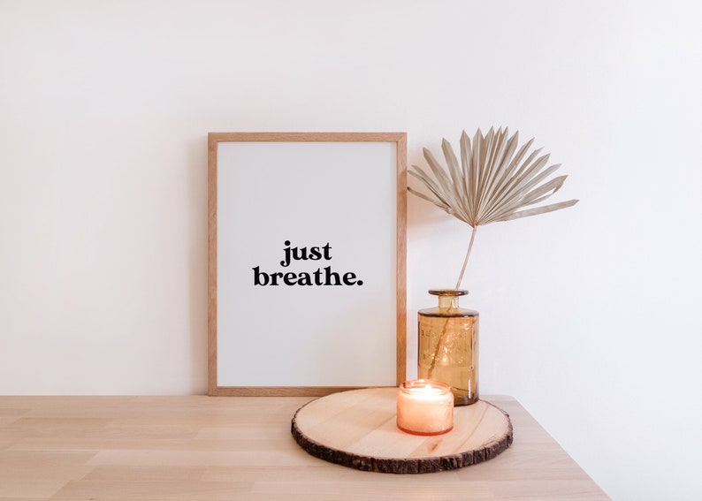 May include: A framed print with the text "just breathe." on a white background. The print is sitting on a wooden table next to a vase with a dried palm leaf and a lit candle.