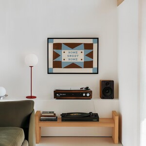 Puede incluir: Una l&aacute;mina de arte mural rectangular en un marco negro con un patr&oacute;n geom&eacute;trico de estrella en marr&oacute;n y azul claro con el texto HOME SWEET HOME centrado en el medio. La l&aacute;mina se muestra en una pared blanca sobre un banco de madera.