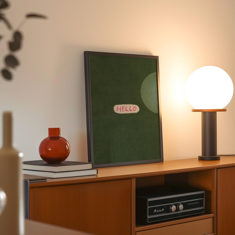 Puede incluir: Una escena interior de mediados de siglo con una obra de arte verde enmarcada con la palabra "HELLO", un jarr&oacute;n naranja redondo, una l&aacute;mpara de globo brillante y un equipo de m&uacute;sica vintage en un mueble de madera. Luz c&aacute;lida.