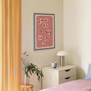 May include: A framed art print with the phrase "Snug as a bug in a rug" in red letters on a pink background. The artwork hangs on a cream-colored wall above a nightstand with a lamp and a blue mug.